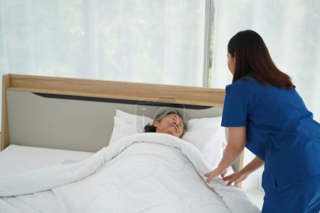 Mitfühlende Krankenschwester, die einer im Bett liegenden Seniorin sanfte Pflege anbietet. Junge Pflegekraft sorgt für Geborgenheit und Wärme der älteren Dame und deckt sie mit weicher Decke für erholsamen Schlaf zu Hause.