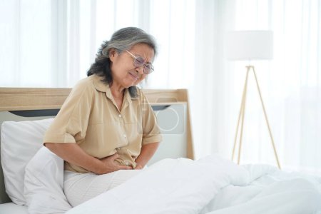 Asiatische Reife Frau mit Brille sitzt auf dem Bett und hält ihren Bauch mit Bauchschmerzen, ihr Gesichtsausdruck voller Schmerzen, Kopierraum