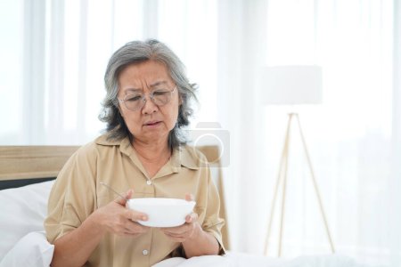 Foto de Asiática Senior mujer con gafas sentado en la cama en la bodega del dormitorio y mirando un tazón de comida, expresión facial emoción con sensación anoréxica - Imagen libre de derechos