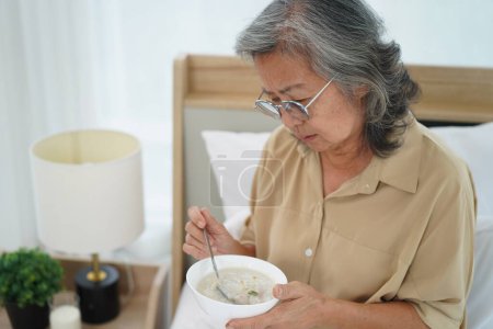 Téléchargez les photos : Asiatique Senior femme portant des lunettes assis sur le lit dans la chambre à coucher tenir et à la recherche d'un bol de repas, expression émotion faciale avec sentiment anorexique - en image libre de droit