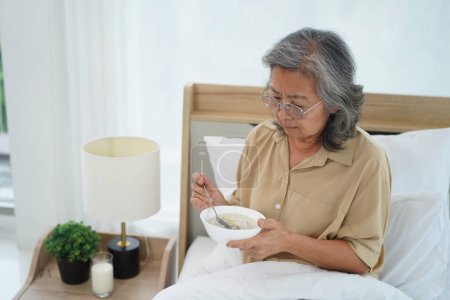 Téléchargez les photos : Asiatique Senior femme portant des lunettes assis sur le lit dans la chambre à coucher tenir et à la recherche d'un bol de repas, expression émotion faciale avec sentiment anorexique - en image libre de droit