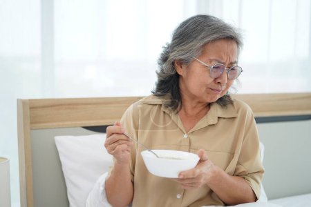 Foto de Asiática Senior mujer con gafas sentado en la cama en el dormitorio celebrar un tazón de comida, expresión facial emoción con sensación anoréxica - Imagen libre de derechos