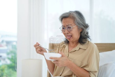 Photo for Asian Senior woman wearing glasses sitting on bed in bedroom hold and looking a bowl of meal, expression facial emotion with feeling anorexic and distasteful - Royalty Free Image