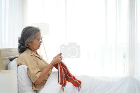 Photo for Asian senior woman sitting on bed in bedroom and crocheting in a handicraft as a hobby or occupational therapy at home, copy space - Royalty Free Image