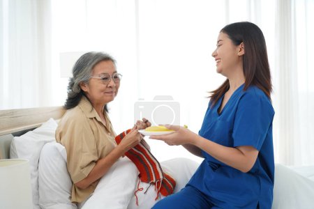 Photo for Asian senior woman sitting on bed in bedroom and crocheting in a handicraft as a hobby or occupational therapy, Young female caregiver give fresh fruit for her to eating at home or nursing home - Royalty Free Image