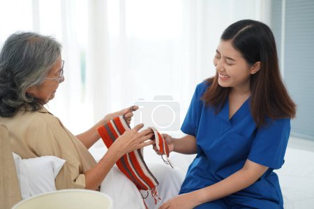 Photo for Asian senior woman sitting on bed in bedroom and crocheting in a handicraft as a hobby or occupational therapy, She teaching to Young female caregiver, They have fun together at home or nursing home - Royalty Free Image