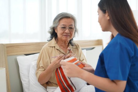 Photo for Selective focus, Asian senior woman sitting on bed in bedroom and crocheting in a handicraft as a hobby or occupational therapy, She smile and talk with Young female caregiver at home or nursing hom - Royalty Free Image