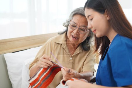 Photo for Selective focus, Asian senior woman sitting on bed in bedroom and crocheting in a handicraft as a hobby or occupational therapy, She smile and talk with Young female caregiver at home or nursing hom - Royalty Free Image