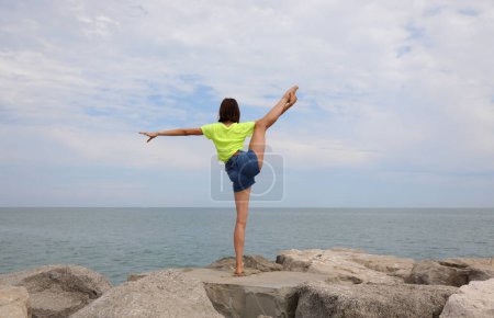 Photo for Young girl does gymnastic exercises to train her balance by straining on one leg on the rocks by the sea - Royalty Free Image