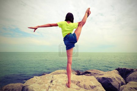 Téléchargez les photos : Jeune fille effectue des exercices de gymnastique sur les rochers au bord de la mer avec effet tonique antique - en image libre de droit