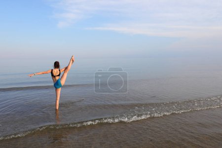 Photo for Slender athletic girl performs gymnastic exercises on the sea near the shore in summer - Royalty Free Image