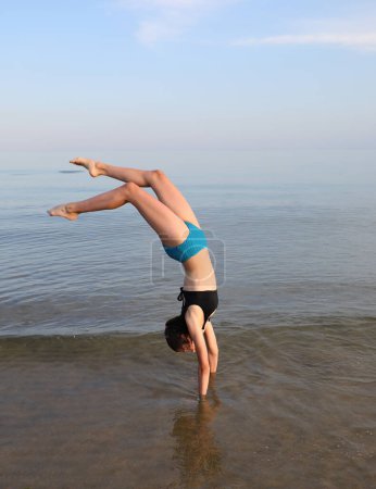 Téléchargez les photos : Young girl carries out gymnastic heads with her head down the sea near the coast - en image libre de droit