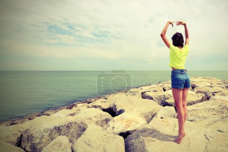 Téléchargez les photos : Jeune fille effectue des exercices de gymnastique sur les rochers au bord de la mer avec effet tonique antique - en image libre de droit