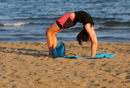 Photo for Young girl training by the sea in summer on the gym mat on the beach at sunset - Royalty Free Image