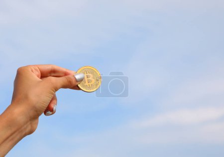 Téléchargez les photos : Main de la jeune fille tenant pièce d'or avec lettre majuscule B symbolisant crypto-monnaie Bitcoin et fond bleu ciel - en image libre de droit