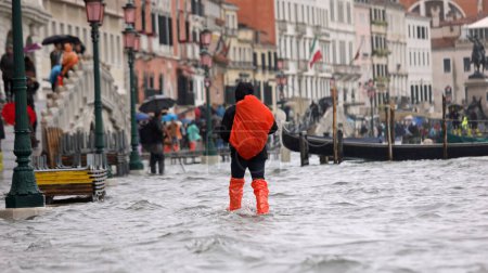 Photo for High tide in Venice Island in Italy and tourist with red backpack and waterproof leggings in the middle of the water - Royalty Free Image