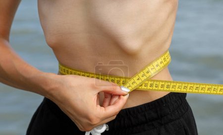 Foto de Niña con trastorno alimenticio llama anorexia mientras mide su abdomen con cinta métrica amarilla - Imagen libre de derechos