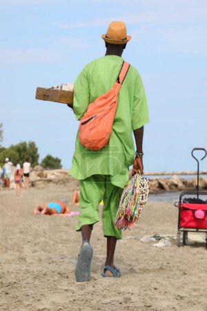 Téléchargez les photos : Vendeur de rue africaine marche sur la plage vendant des marchandises avec robe verte - en image libre de droit