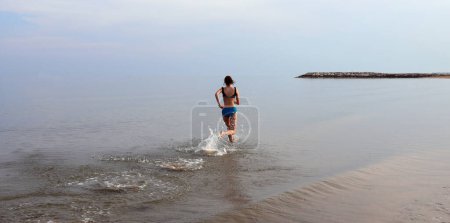 Photo for Slender athletic girl runs with wide strides on the sea near the shore in summer - Royalty Free Image