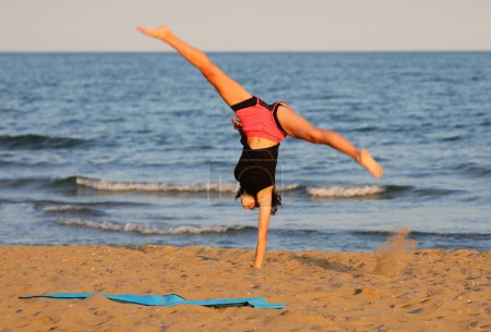 Foto de Atlética chica delgada por el mar hace entrenamiento gimnástico con la cabeza hacia abajo para fortalecer los músculos - Imagen libre de derechos