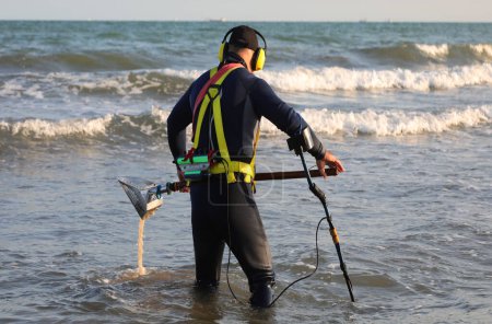 Téléchargez les photos : Récupérateur à la recherche d'objets métalliques perdus avec détecteur de métaux au milieu de l'eau de mer le soir - en image libre de droit