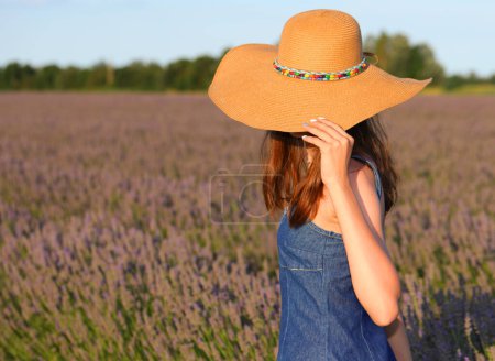 Photo for Young slender top model girl in jeans dress and miniskirt and straw hat among lavender flowers at sunset - Royalty Free Image
