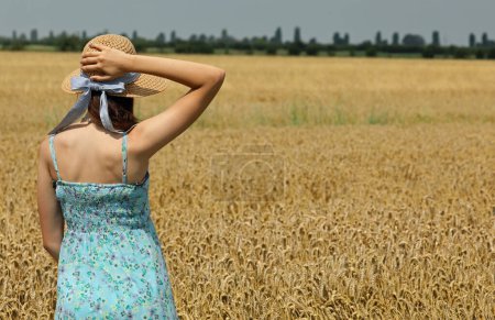 Foto de Muchacha joven con sombrero de paja mira el campo de trigo maduro en verano - Imagen libre de derechos