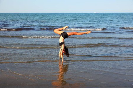 Foto de Joven chica realiza la pierna se divide durante el entrenamiento en la playa por mar - Imagen libre de derechos