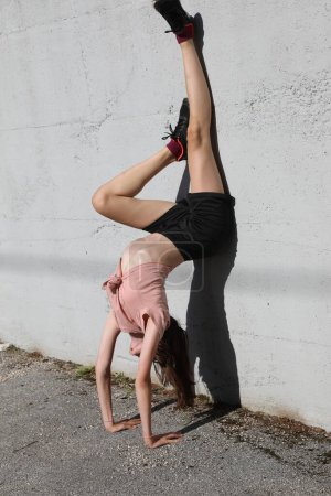 Téléchargez les photos : Jeune fille faisant des exercices assis sur le mur avec ses jambes pointant vers le haut et les mains sur le sol - en image libre de droit