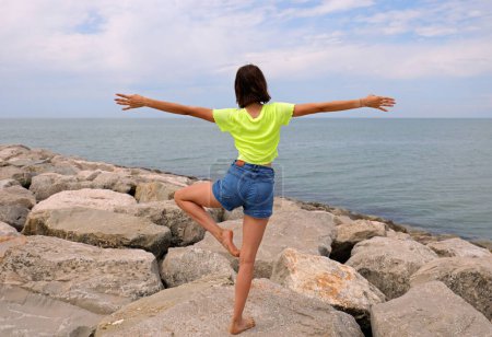 Photo for Young slender girl performs gymnastic exercises on the rocks by the sea in jeans shorts and short-sleeved t-shirt in summer - Royalty Free Image