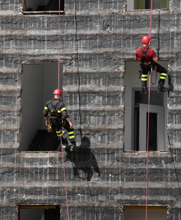 Foto de Dos valientes bomberos en arnés suben a un edificio destruido con cuerda y casco rojo - Imagen libre de derechos