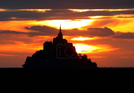 Foto de Encantadora puesta de sol con cielo ardiente con nubes anaranjadas y rojas y la silueta de la antigua abadía del Mont Saint Michel en el norte de Francia - Imagen libre de derechos