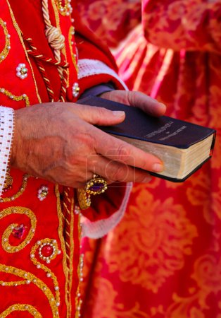 Foto de Biblia con las Sagradas Escrituras en las manos del sacerdote con sotana roja durante la ceremonia religiosa en la iglesia - Imagen libre de derechos