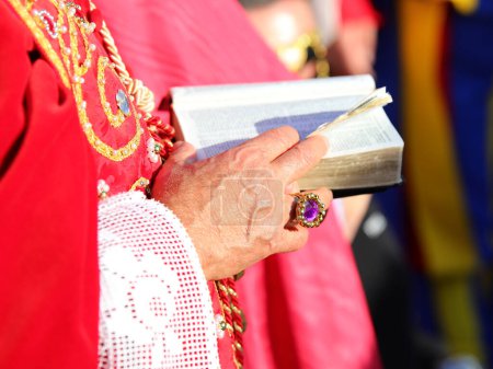 älterer Bischof mit roter Soutane und der alten Bibel mit den Heiligen Schriften in der Hand mit einem Ring mit rotem Rubin während des religiösen Ritus