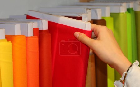 Téléchargez les photos : Main d'une jeune fille choisissant tissu de couleur rouge dans la mercerie avec de nombreux tissus colorés - en image libre de droit