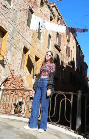 Téléchargez les photos : Jeune fille avec masque vénitien portant un pantalon en jeans bleu posant près des maisons avec des vêtements traînant au soleil en Italie - en image libre de droit