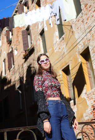 young girl with venetian mask wearing blue jeans trousers posing  near houses with clothes hanging