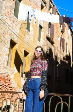 Mädchen mit venezianischer Maske trägt Jeanshose und posiert in der Nähe von Häusern mit Kleidung, die zum Trocknen in der Sonne in Venedig in Italien hängt
