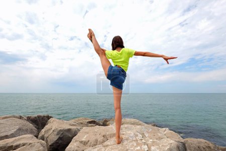 chica joven realiza ejercicios de gimnasia sobre rocas por mar con pantalones vaqueros cortos en verano