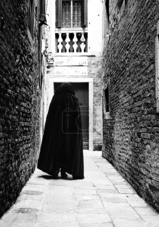 personne à capuche avec manteau foncé marchant dans une ruelle étroite de la ville avec un effet dramatique en noir et blanc