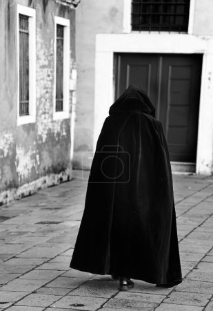 hooded anonymous person walking wearing an old black tabard as a cloak on the narrow alley of the city center