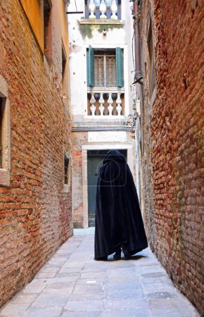Photo for Anonymous figure in a hood walks through a narrow city alley, wearing a worn black tabard as a cloak - Royalty Free Image