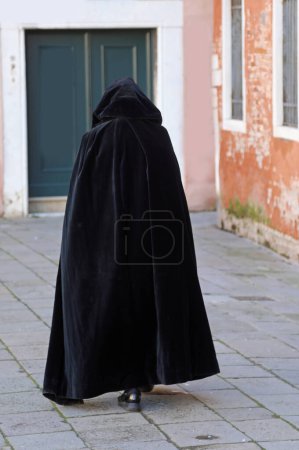 figure in a hood walks through a city alley wearing a worn black tabard as a cloak