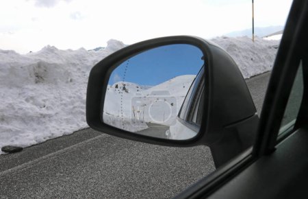 Rearview mirror of the passenger car on the mountain road in winter with snow and reflection of the past