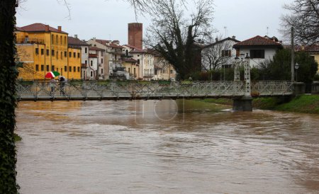 Raging river during flooding with risk of overflow and nearly submerged pedestrian iron bridge