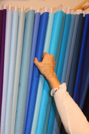 Téléchargez les photos : Main d'une jeune fille choisissant un tissu de couleur bleu profond dans la mercerie avec de nombreux tissus colorés - en image libre de droit