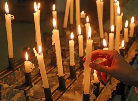 weibliche Hand mit einer brennenden Kerze in einer Kirche inmitten eines flackernden Flammenmeeres aus Kerzen frommer Pilger