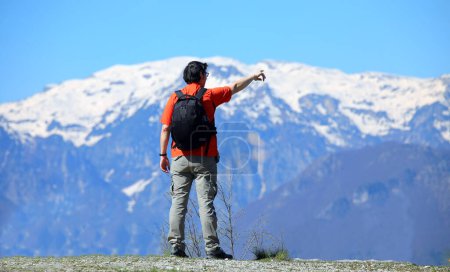young Man hiker with black backpack standing on a mountain trail looking at snowcapped peaks