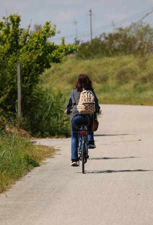 woman with a backpack on their shoulders pedaling leisurely on the cycle path in the countryside near an embankment