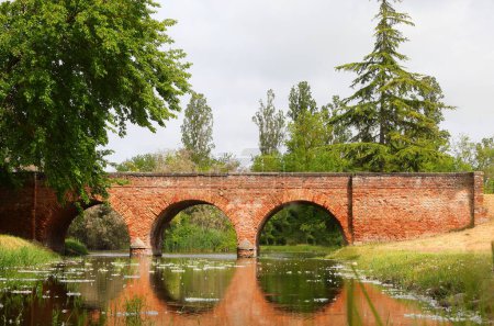 Ancient red brick bridge with visible masonry and the reflection of its three arches on the water of a calm river near a forest